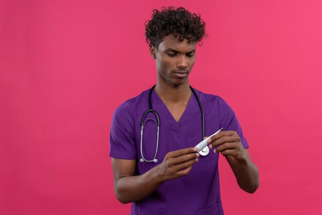 Un confundido joven apuesto médico de piel oscura con cabello rizado vistiendo uniforme violeta con estetoscopio mirando el termómetro