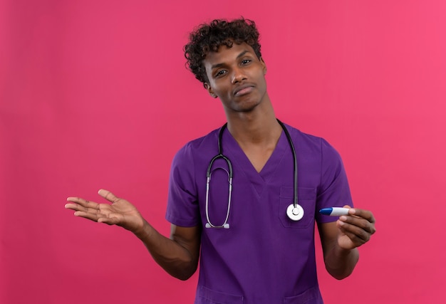 Un confundido joven apuesto médico de piel oscura con cabello rizado vistiendo uniforme violeta con estetoscopio sosteniendo termómetro