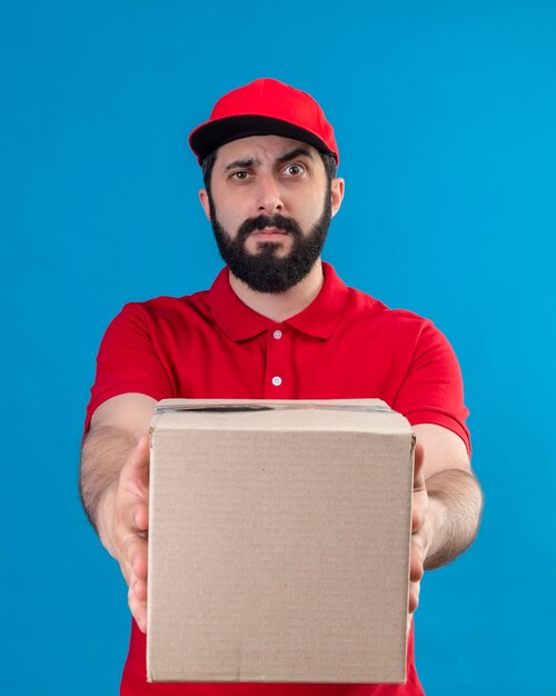 Confundido joven apuesto repartidor caucásico vestido con uniforme rojo y gorra estirando la caja de cartón hacia la cámara aislada en azul