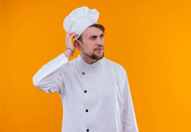 Un confundido joven chef barbudo con uniforme blanco sosteniendo la mano en la cabeza mientras piensa en una pared naranja