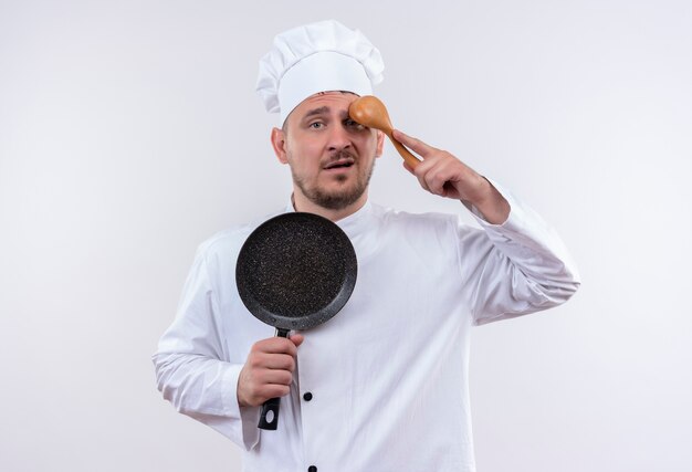 Confundido joven cocinero guapo en uniforme de chef sosteniendo una sartén y poniendo una cuchara en la frente aislada en la pared blanca