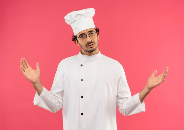 Confundido joven cocinero vistiendo uniforme de chef y gafas extiende las manos