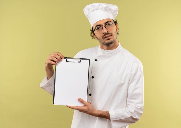 Confundido joven cocinero vistiendo uniforme de chef y gafas sosteniendo el portapapeles