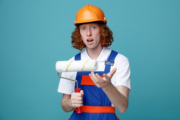 Confundido joven constructor hombre en uniforme sosteniendo cepillo de rodillos aislado sobre fondo azul.