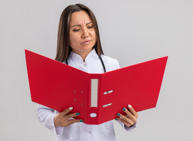 Confundido joven doctora asiática vistiendo bata médica y estetoscopio sosteniendo y mirando la carpeta abierta aislada en la pared blanca