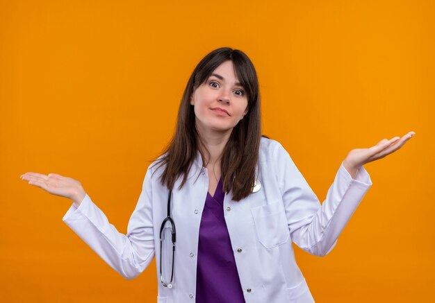 Confundido joven doctora en bata médica con estetoscopio mantiene ambas manos abiertas sobre fondo naranja aislado con espacio de copia