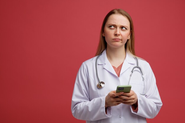 Confundido joven doctora rubia vistiendo bata médica y estetoscopio alrededor del cuello con teléfono móvil mirando hacia arriba con los labios fruncidos