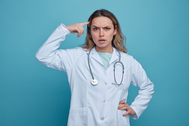 Confundido joven doctora vistiendo bata médica y estetoscopio alrededor del cuello manteniendo la mano en la cintura haciendo gesto de pensar