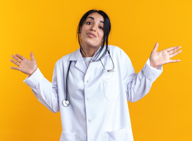 Confundido joven doctora vistiendo bata médica con estetoscopio extendiendo las manos aisladas en la pared amarilla