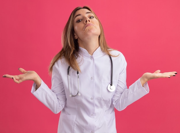Confundido joven doctora vistiendo bata médica con estetoscopio extendiendo las manos aisladas en la pared rosa