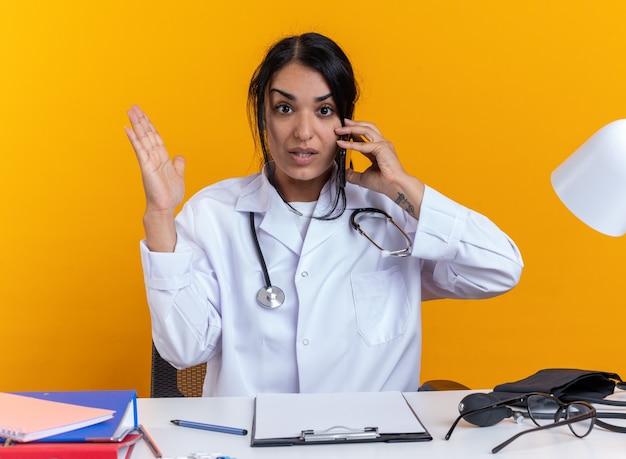 Foto gratuita confundido joven doctora vistiendo bata médica con estetoscopio se sienta a la mesa con herramientas médicas habla por teléfono aislado en la pared amarilla