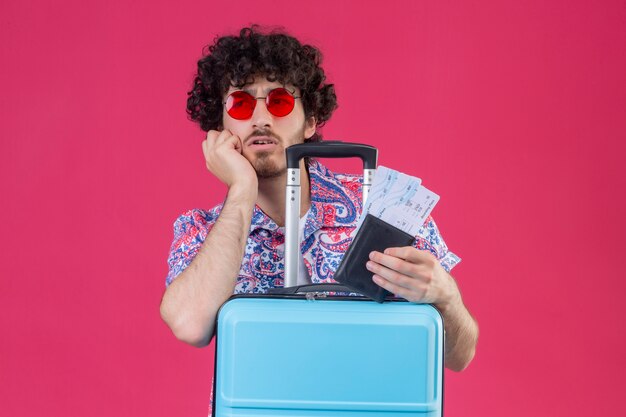 Confundido joven guapo viajero rizado hombre con gafas de sol sosteniendo billetera y boletos de avión poniendo la mano en la mejilla con la maleta en el espacio rosa aislado con espacio de copia