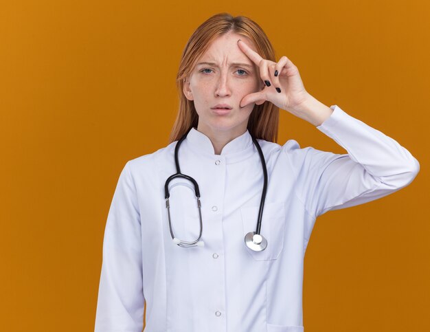 Confundido joven médico de jengibre vistiendo bata médica y estetoscopio tocando la cara