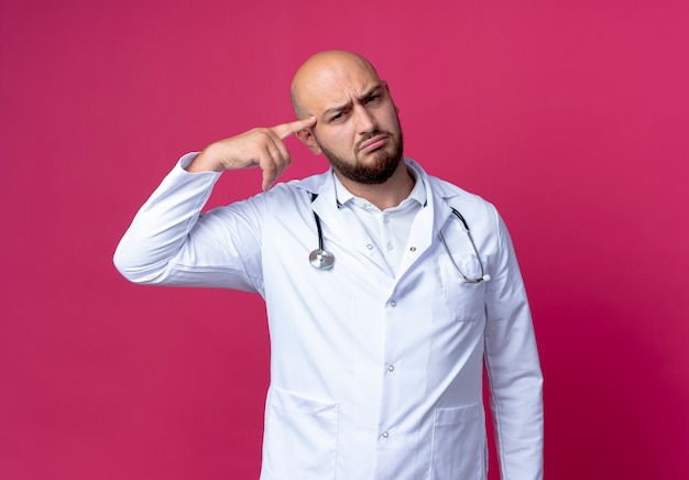 Confundido joven médico varón calvo vistiendo bata médica y estetoscopio poniendo el dedo en la frente aislado en la pared rosa