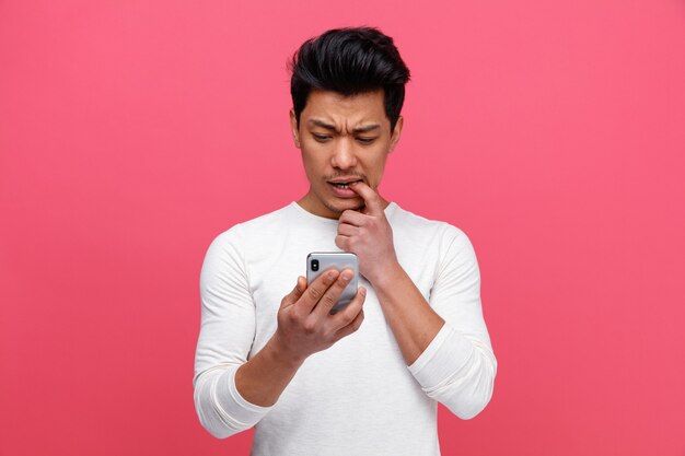 Confundido joven mordiendo el dedo sosteniendo y mirando el teléfono móvil