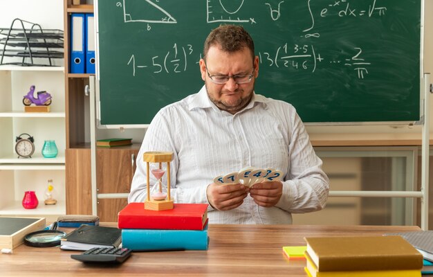 Confundido joven profesor con gafas sentado en el escritorio con útiles escolares en el aula sosteniendo y mirando el número de ventiladores