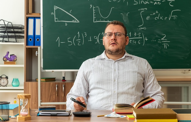 Confundido joven profesor rubio con gafas sentado en el escritorio con herramientas escolares en el aula sosteniendo lupa agarrando libro abierto mirando a la cámara