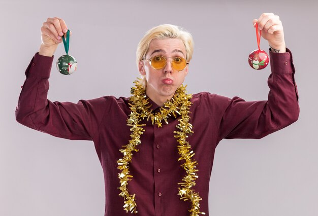 Confundido joven rubio con gafas con guirnalda de oropel alrededor del cuello levantando adornos navideños mirando a cámara aislada sobre fondo blanco