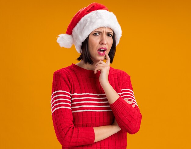 Confundido joven vistiendo gorro de Papá Noel mirando a la cámara poniendo figer en el labio aislado sobre fondo naranja con espacio de copia