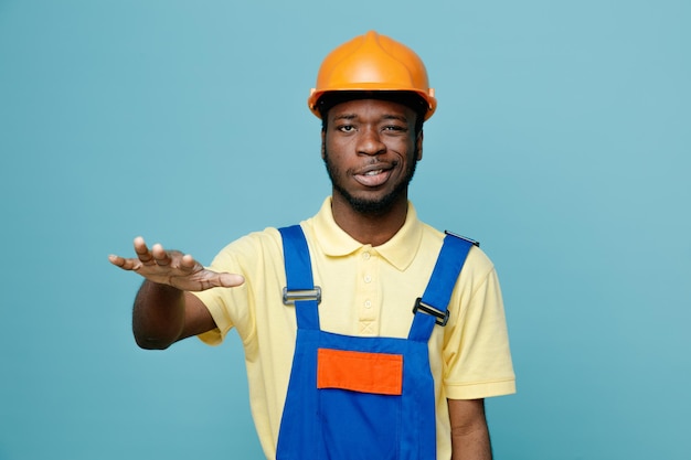 Confundido mano levantada joven constructor afroamericano en uniforme aislado sobre fondo azul.
