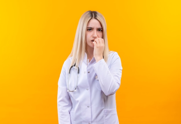 Confundido médico joven con estetoscopio en bata médica puso su mano en la barbilla en la pared amarilla aislada