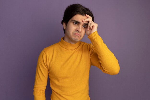 Confundido mirando al lado chico guapo joven con suéter de cuello alto amarillo poniendo la mano en el templo aislado en la pared púrpura