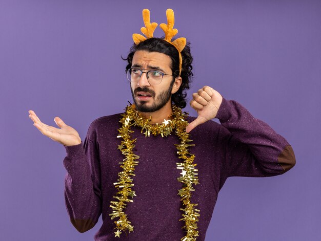 Confundido mirando al lado joven guapo con aro de pelo de navidad con guirnalda en el cuello mostrando los puntos del pulgar hacia abajo con la mano al lado aislado sobre fondo azul con espacio de copia