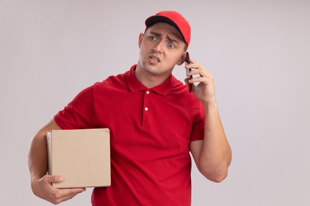 Confundido mirando al lado joven repartidor vestido con uniforme con gorra sosteniendo la caja habla por teléfono aislado en la pared blanca con espacio de copia