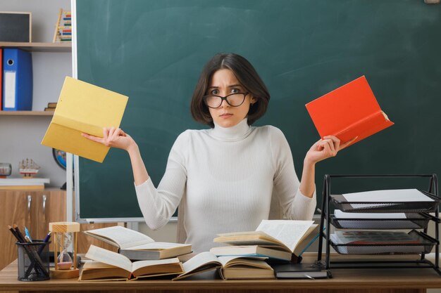 confundido mirando a la cámara joven maestra con gafas sosteniendo libros sentados en el escritorio con herramientas escolares en el aula