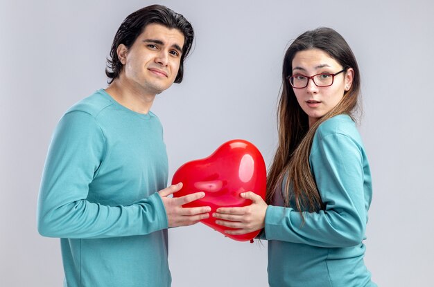 Confundido mirando a la cámara pareja joven en el día de San Valentín sosteniendo globo corazón aislado sobre fondo blanco.