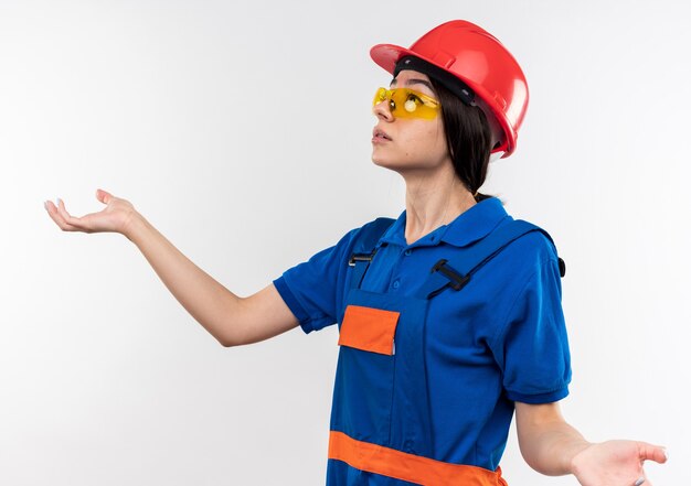 Confundido mirando a la mujer joven constructor de lado en uniforme con gafas extendiendo las manos