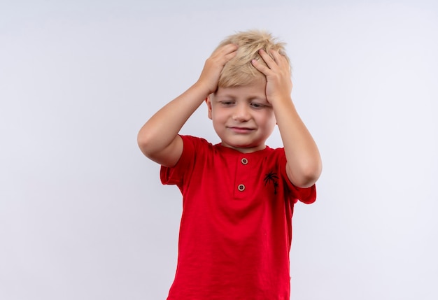 Un confundido niño rubio lindo en camiseta roja sosteniendo la cabeza entre las manos mientras mira hacia abajo en una pared blanca