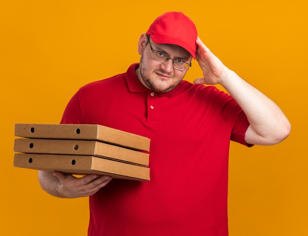 Confundido repartidor joven con sobrepeso en gafas ópticas sosteniendo cajas de pizza y poniendo la mano en la cabeza aislada en la pared naranja con espacio de copia