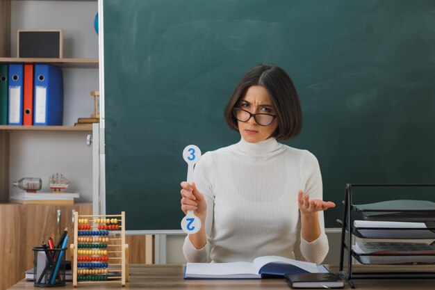 confundido sosteniendo la mano hacia la cámara joven maestra con anteojos sosteniendo un ventilador sentado en el escritorio con herramientas escolares en el aula
