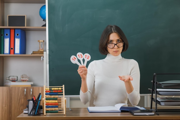 confundido sosteniendo la mano hacia la cámara joven maestra con anteojos sosteniendo un ventilador sentado en el escritorio con herramientas escolares en el aula