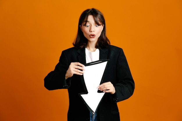 Confundido sosteniendo marca de dirección joven hermosa mujer vistiendo chaqueta negra aislado sobre fondo naranja