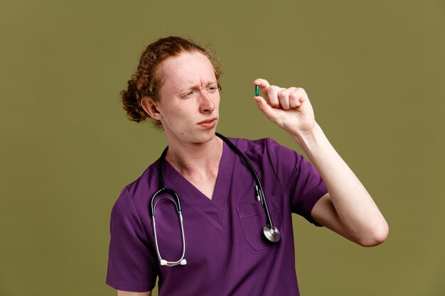 Confundido sosteniendo píldoras joven médico masculino vistiendo uniforme con estetoscopio aislado sobre fondo verde