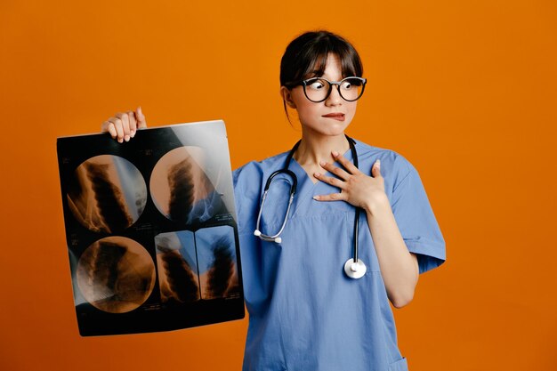 Confundido sosteniendo rayos X joven doctora vistiendo uniforme fith estetoscopio aislado sobre fondo naranja