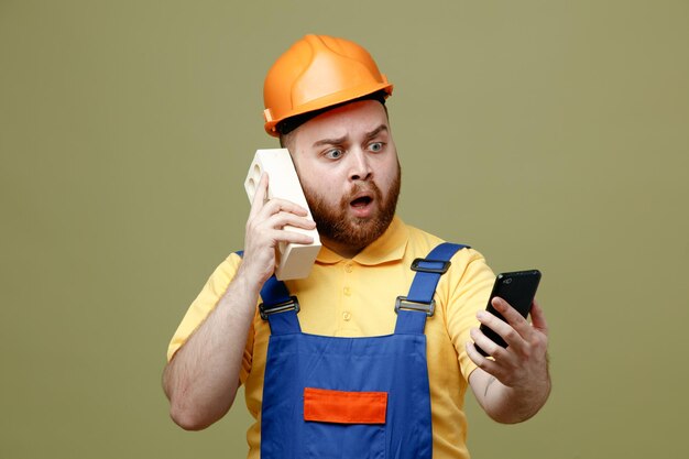 Confundido sosteniendo teléfono poniendo ladrillo en la cabeza joven constructor hombre en uniforme aislado sobre fondo verde