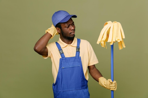 confundido sosteniendo un trapeador joven limpiador afroamericano en uniforme con guantes aislados en fondo verde