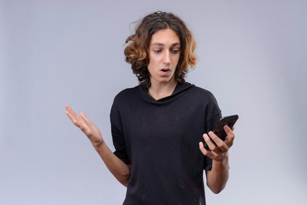 Confundir al chico con el pelo largo en camiseta negra sosteniendo un teléfono en la pared blanca