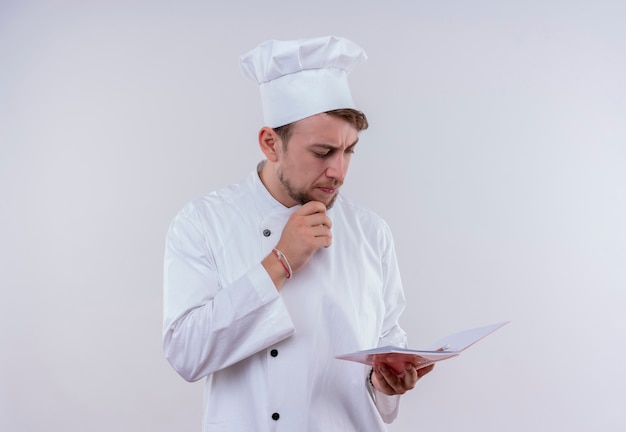 Un confuso joven chef barbudo vestido con uniforme de cocina blanco y sombrero pensando y mirando el cuaderno en una pared blanca