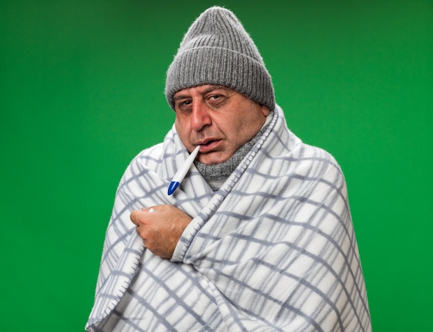 Congelación adulto enfermo hombre caucásico con bufanda alrededor del cuello con gorro de invierno envuelto en plaid sosteniendo el termómetro en la boca aislado en la pared verde con espacio de copia