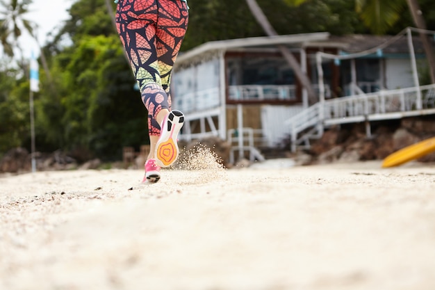 Congelar la toma de acción de la mujer jogger en ropa deportiva corriendo en la playa de arena en un día soleado.