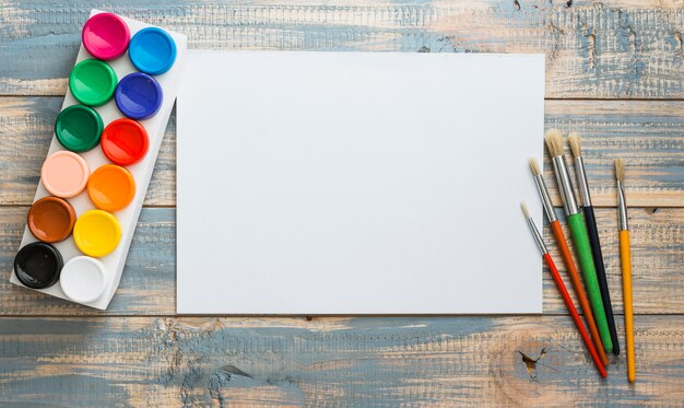 Conjunto de acuarelas y pincel con papel blanco vacío en blanco sobre mesa de madera vieja