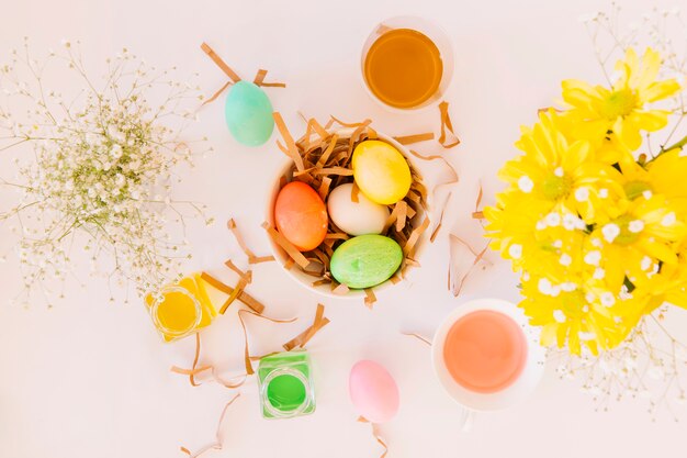 Conjunto de brillantes huevos de Pascua en un tazón entre flores frescas y latas de líquido colorante