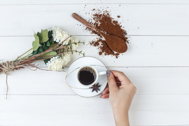 Conjunto de café molido, flores, especias y mano femenina sosteniendo una taza de café sobre un fondo de madera. endecha plana.