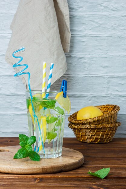 Conjunto de canasta con limones, paño blanco, tabla de cortar y vaso de jugo de limón sobre una superficie de madera y blanca. vista lateral.