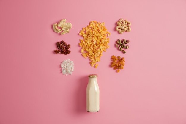 Conjunto de cereales, pistacho, pasas, nueces pecanas, manzana seca, anacardo, coco alrededor de una botella de leche aislada sobre fondo rosa. Desayuno nutritivo rico en vitaminas para consumir, concepto de nutrición.