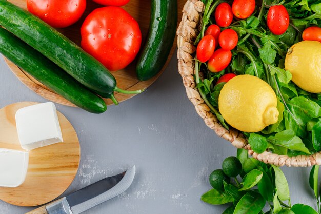 Conjunto de cuchillo, limón, pepino, queso, verduras y tomates en una tabla de cortar sobre una superficie gris